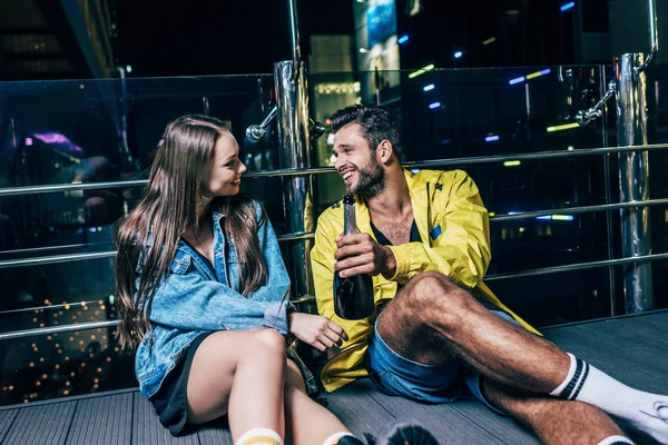 Guapo novio con botella y atractiva novia sonriendo y hablando en la noche de la ciudad - foto de stock