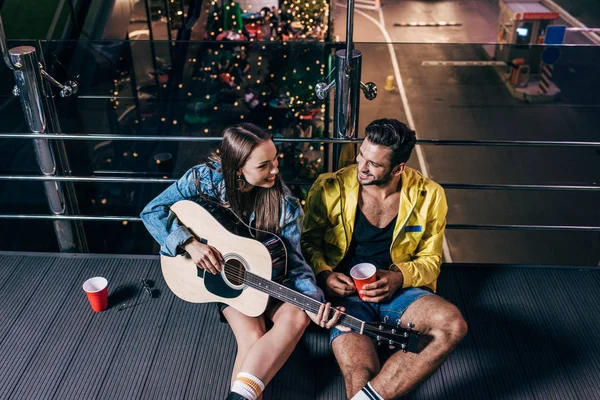 Vista de ángulo alto de novio guapo con taza de plástico y novia atractiva tocando la guitarra acústica en la ciudad de la noche - foto de stock