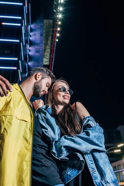 Low angle view of handsome boyfriend and attractive girlfriend smiling and hugging in night city — Stock Photo