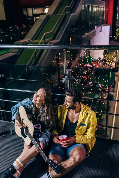 Vista ad alto angolo di bel ragazzo con tazza di plastica e fidanzata attraente suonare la chitarra acustica nella città di notte — Foto stock