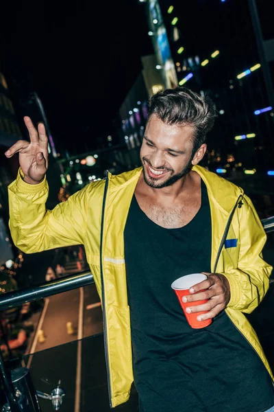 Handsome man in yellow jacket showing peace sign and holding plastic cup in night city — Stock Photo