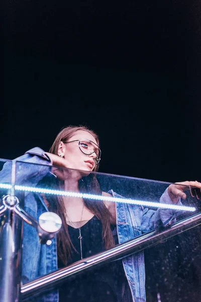 Attraente donna in giacca di denim e occhiali guardando lontano in città notte — Foto stock