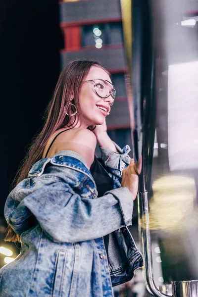 Attraktive und lächelnde Frau in Jeansjacke und Brille, die nachts in der Stadt wegschaut — Stockfoto