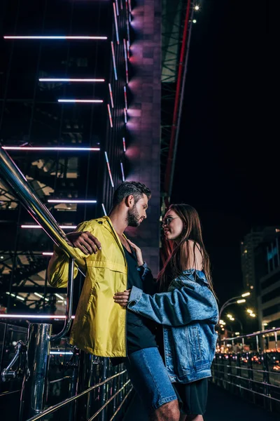 Side view of handsome boyfriend and attractive girlfriend in denim jacket hugging in night city — Stock Photo