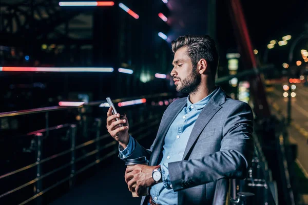 Bell'uomo d'affari in abbigliamento formale utilizzando smartphone e tenendo tazza di carta nella città di notte — Foto stock