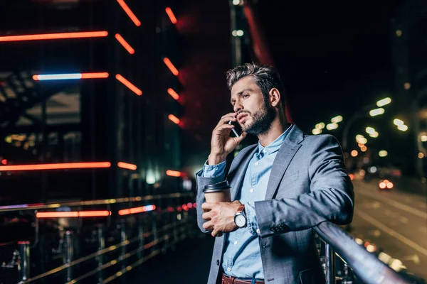 Schöner Geschäftsmann in festlicher Kleidung, der mit dem Smartphone spricht und in der nächtlichen Stadt Pappbecher in der Hand hält — Stockfoto