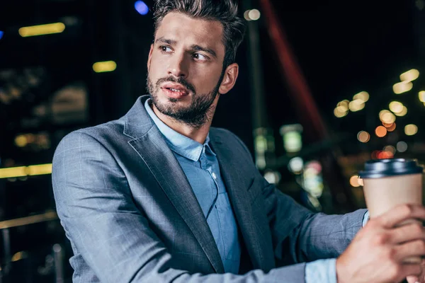 Guapo hombre de negocios en ropa formal sosteniendo taza de papel en la ciudad de la noche - foto de stock