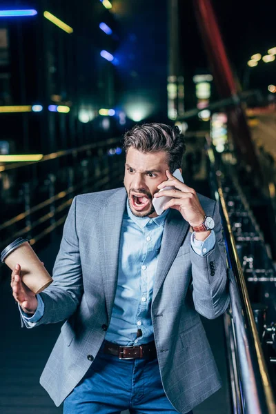 Wütender gutaussehender Geschäftsmann in festlicher Kleidung, der mit dem Smartphone spricht und in der nächtlichen Stadt Pappbecher in der Hand hält — Stockfoto