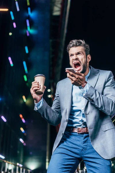 Angry handsome businessman in formal wear talking on smartphone and holding paper cup in night city — Stock Photo