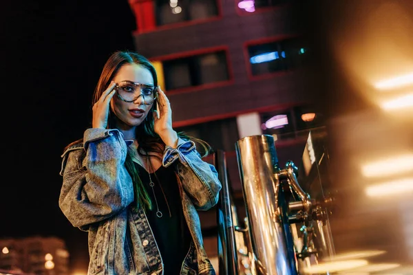 Jolie femme en jean veste et lunettes regardant la caméra dans la ville de nuit — Photo de stock