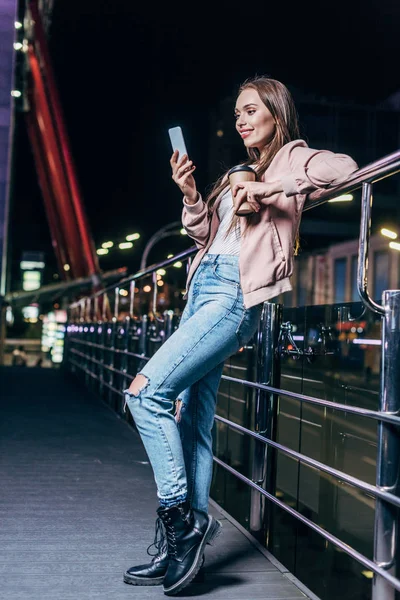 Mujer sonriente con chaqueta rosa usando smartphone y sosteniendo taza de papel en la ciudad nocturna - foto de stock