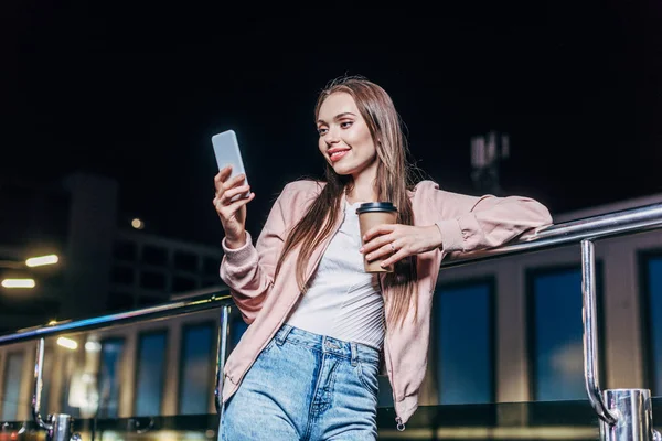 Donna sorridente in giacca rosa utilizzando smartphone e tenendo tazza di carta in città notte — Foto stock
