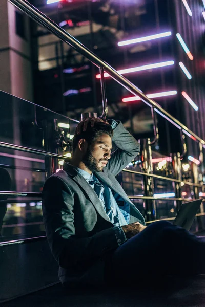 Homem de negócios bonito no desgaste formal sentado e usando laptop na cidade noturna — Fotografia de Stock