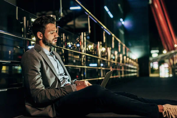 Handsome businessman in formal wear sitting and using laptop in night city — Stock Photo