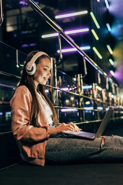 Mujer sonriente en chaqueta rosa con auriculares usando el ordenador portátil en la ciudad de la noche - foto de stock