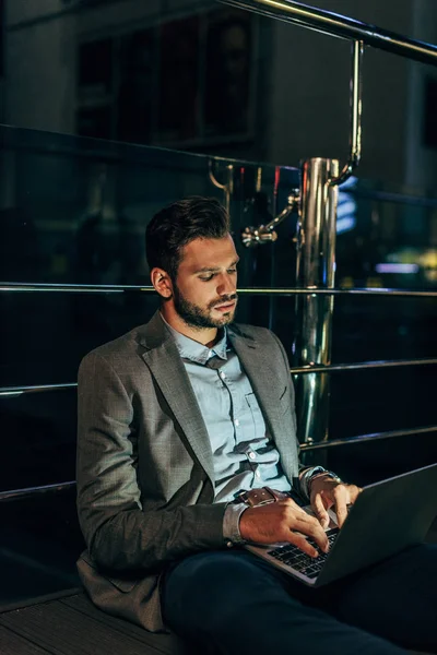 Handsome businessman in formal wear sitting and using laptop in night city — Stock Photo