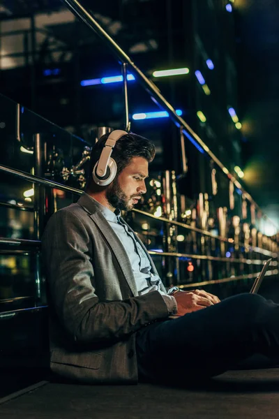 Schöner Geschäftsmann in formeller Kleidung mit Kopfhörern sitzt und Laptop in der Nacht Stadt — Stockfoto