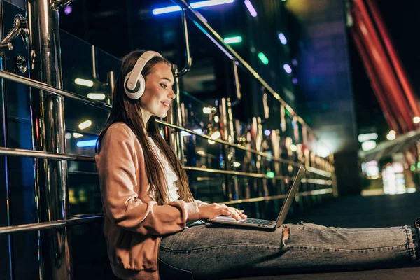 Mulher sorridente em jaqueta rosa com fones de ouvido usando laptop na cidade noturna — Fotografia de Stock