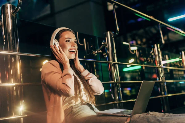 Smiling woman in pink jacket listening music with headphones in night city — Stock Photo