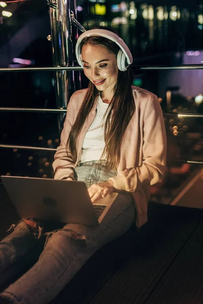 Mujer sonriente en chaqueta rosa con auriculares usando el ordenador portátil en la ciudad de la noche - foto de stock