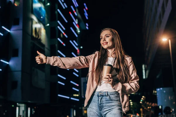 Attractive woman in pink jacket hitching and holding paper cup in night city — Stock Photo