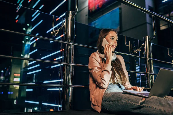 Mujer sonriente en chaqueta rosa con auriculares hablando en el teléfono inteligente en la ciudad de la noche - foto de stock