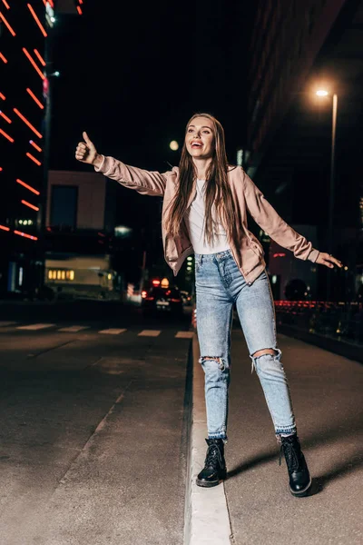 Attractive woman in pink jacket hitching and smiling in night city — Stock Photo