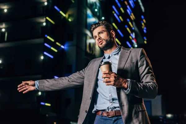 Handsome businessman in formal wear hitching and holding paper cup in night city — Stock Photo