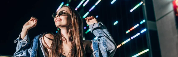 Tiro panorâmico de mulher atraente em jaqueta de ganga e óculos olhando para longe na cidade noturna — Fotografia de Stock