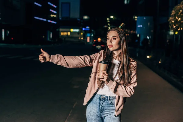 Atractiva mujer en chaqueta rosa enganche y sosteniendo taza de papel en la ciudad de la noche - foto de stock