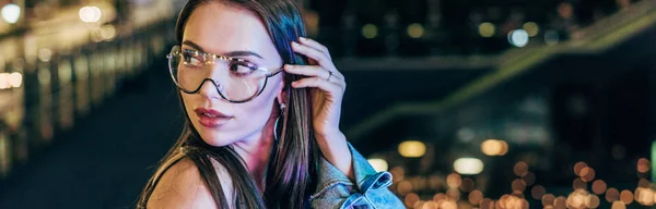 Tiro panorâmico de mulher atraente em óculos olhando para longe na cidade noturna — Fotografia de Stock