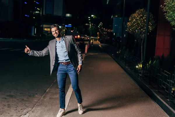 Handsome businessman in formal wear thumbing and smiling in night city — Stock Photo