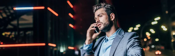 Plano panorámico de hombre de negocios guapo en ropa formal hablando en el teléfono inteligente en la ciudad de noche - foto de stock