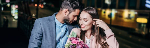 Panoramic shot of handsome businessman giving bouquet and kissing attractive woman in night city — Stock Photo