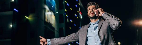 Plano panorámico de hombre de negocios guapo en ropa formal hablando en el teléfono inteligente y pulgar en la ciudad de la noche - foto de stock