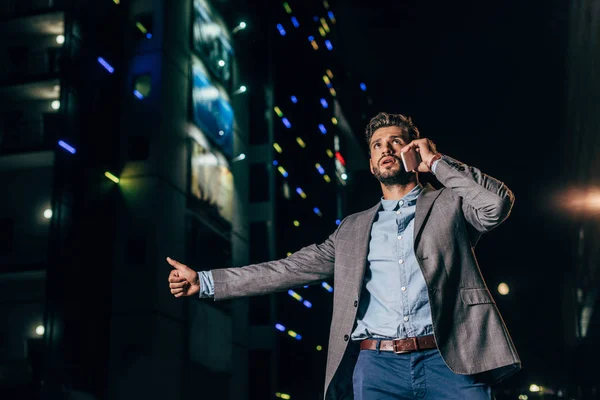 Handsome businessman in formal wear talking on smartphone and thumbing in night city — Stock Photo