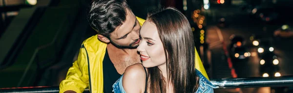 Plan panoramique de petit ami embrassant petite amie attrayante et souriante dans la ville de nuit — Photo de stock