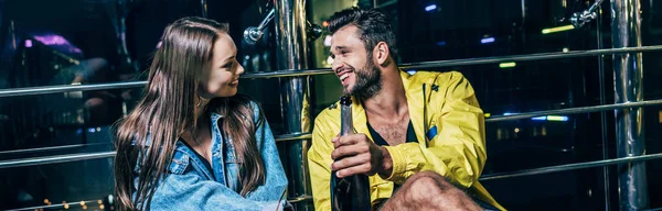 Tiro panorâmico de namorado com garrafa e namorada sorridente falando na cidade noturna — Fotografia de Stock