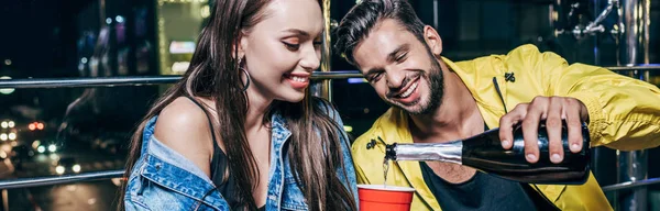 Plan panoramique de beau petit ami souriant versant du champagne à la tasse en plastique dans la ville de nuit — Photo de stock