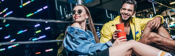 Plano panorámico de novio guapo y novia atractiva con copas de plástico sonriendo en la ciudad de la noche - foto de stock