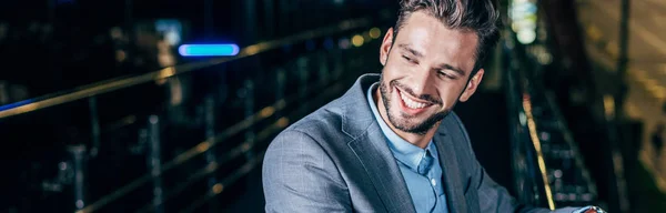 Plano panorámico de hombre de negocios guapo en ropa formal sonriendo y mirando hacia otro lado en la ciudad de la noche - foto de stock