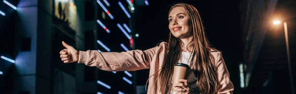 Plano panorámico de mujer atractiva en chaqueta rosa enganche y sosteniendo taza de papel en la ciudad de la noche - foto de stock