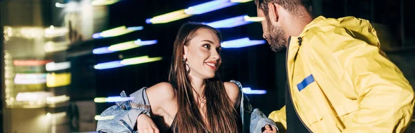 Panoramic shot of smiling girlfriend and boyfriend talking and looking at each other in night city — Stock Photo