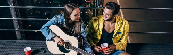 Plano panorámico de novio guapo con taza de plástico y novia atractiva tocando la guitarra acústica en la ciudad de la noche - foto de stock