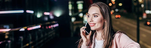 Panoramic shot of attractive woman in pink jacket smiling and talking on smartphone in night city — Stock Photo
