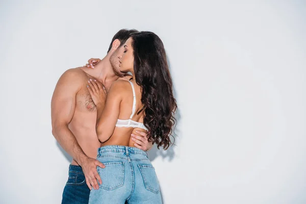 Shirtless man embracing girlfriend in blue jeans and bra on grey background — Stock Photo