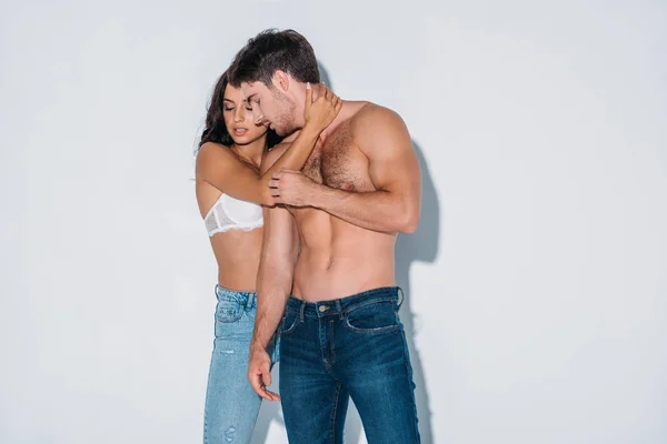 Sexy chica en azul jeans y blanco sujetador abrazando sin camisa novio en gris fondo - foto de stock