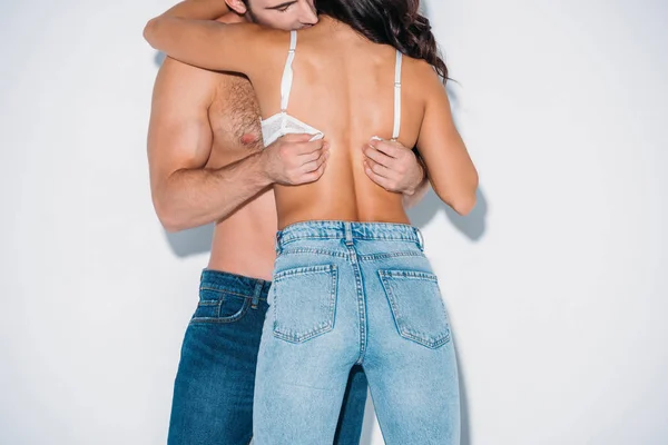 Vue recadrée du jeune homme décoller soutien-gorge de petite amie en jeans bleu sur fond gris — Photo de stock