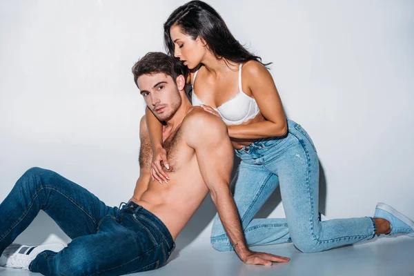 Fille sexy en jeans en soutien-gorge blanc étreignant petit ami assis et regardant la caméra sur fond gris — Photo de stock