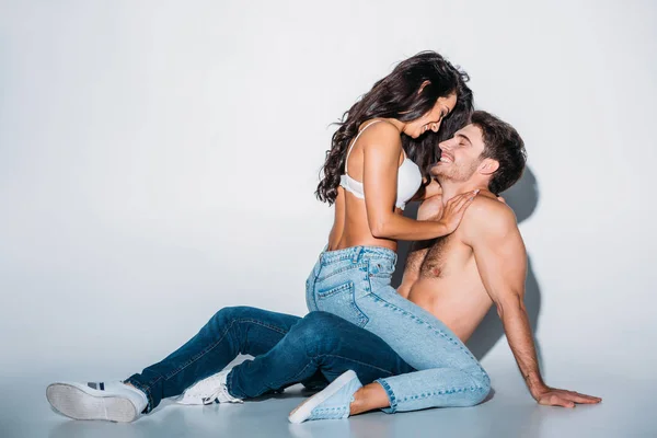 Smiling girl hugging happy shirtless boyfriend sitting on grey background — Stock Photo